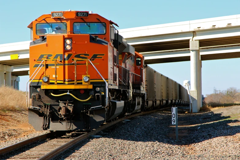 an orange train on a track beneath a white bridge