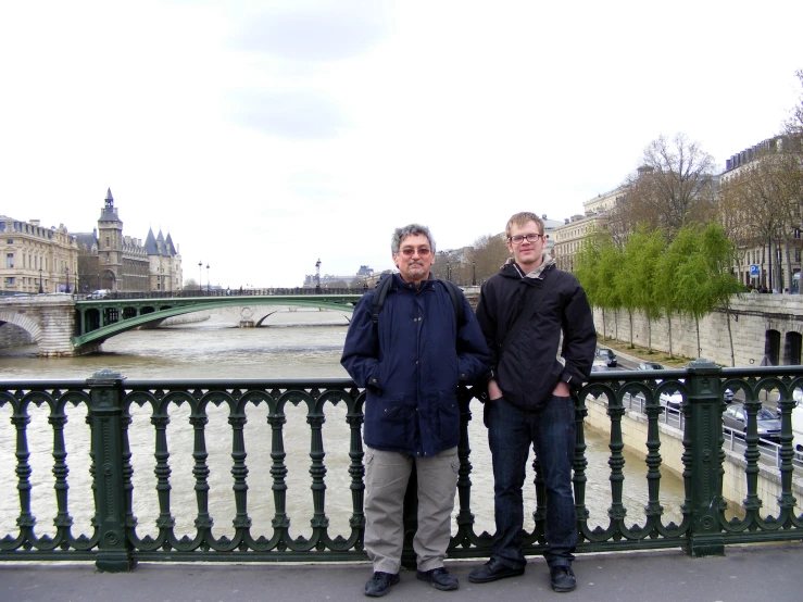 two men standing in front of an iron fence overlooking a river