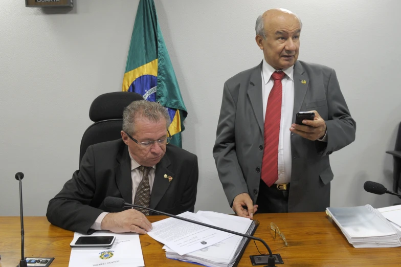 two men in business suits are at a wooden table while looking at a cell phone