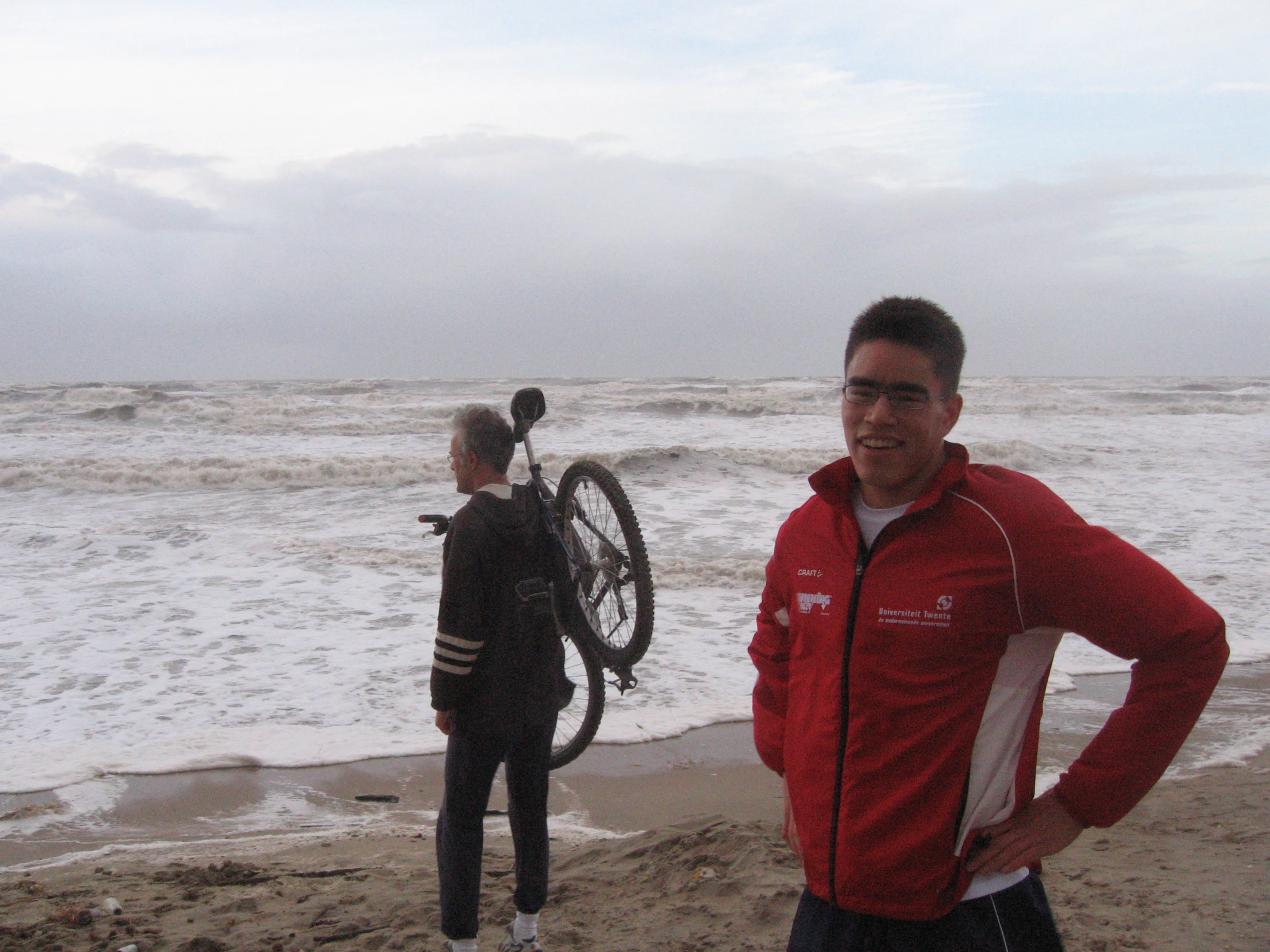 a man standing next to the ocean with another person holding up a bicycle