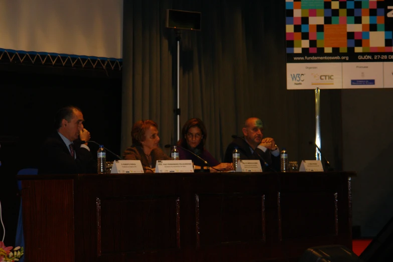 four people sit at the front of a panel, on stage with microphones