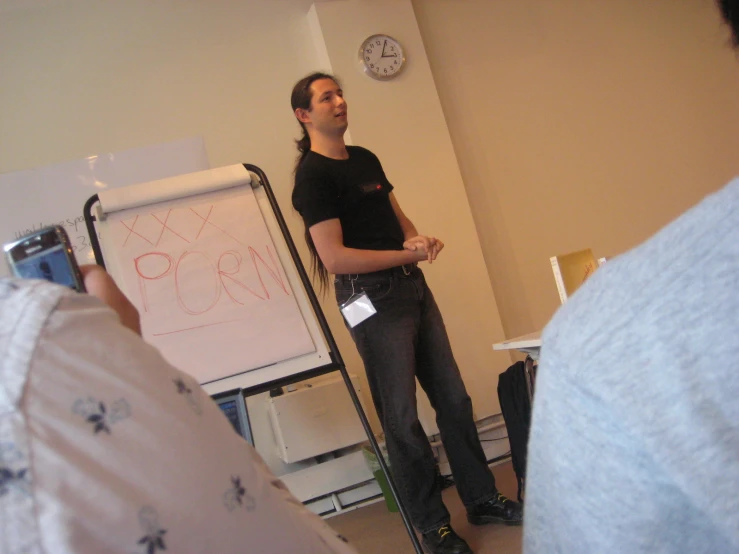 a young man holding a camera stands in front of a classroom and gestures