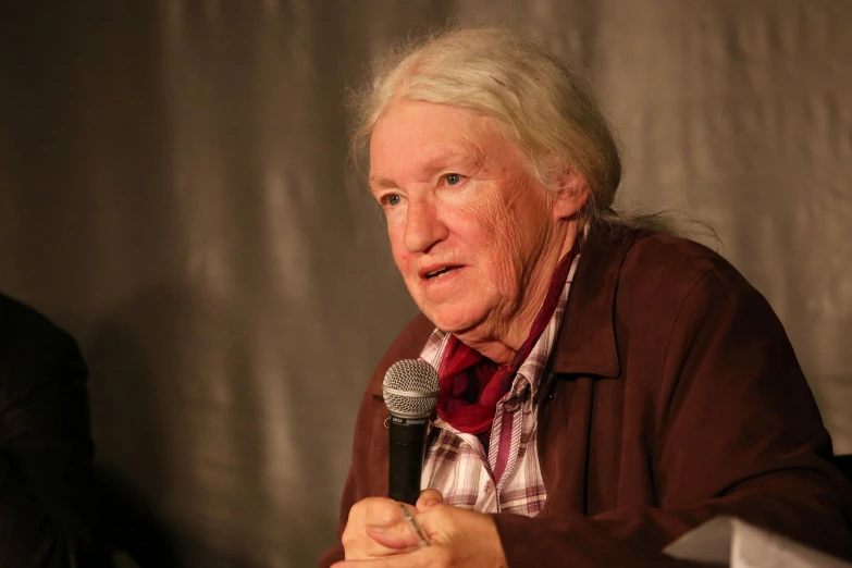 an older woman wearing a brown top and tie holding a microphone in front of her