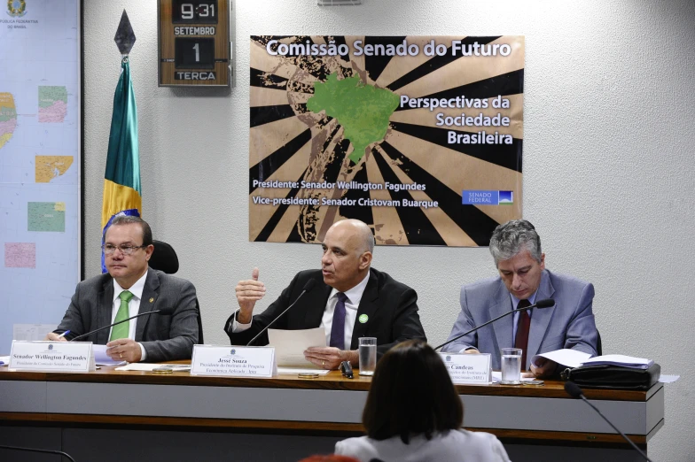 three men sit at a table with papers and microphone