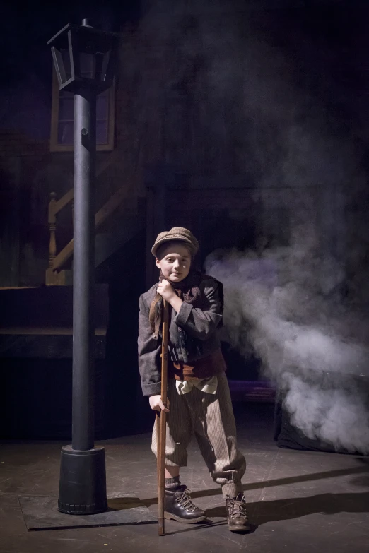 a boy wearing an unoned jacket stands near a pole, smoking a pipe, with a cane and looking sideways