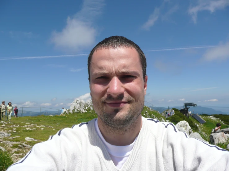 man in white shirt taking a selfie on grassy hill with mountains in the background