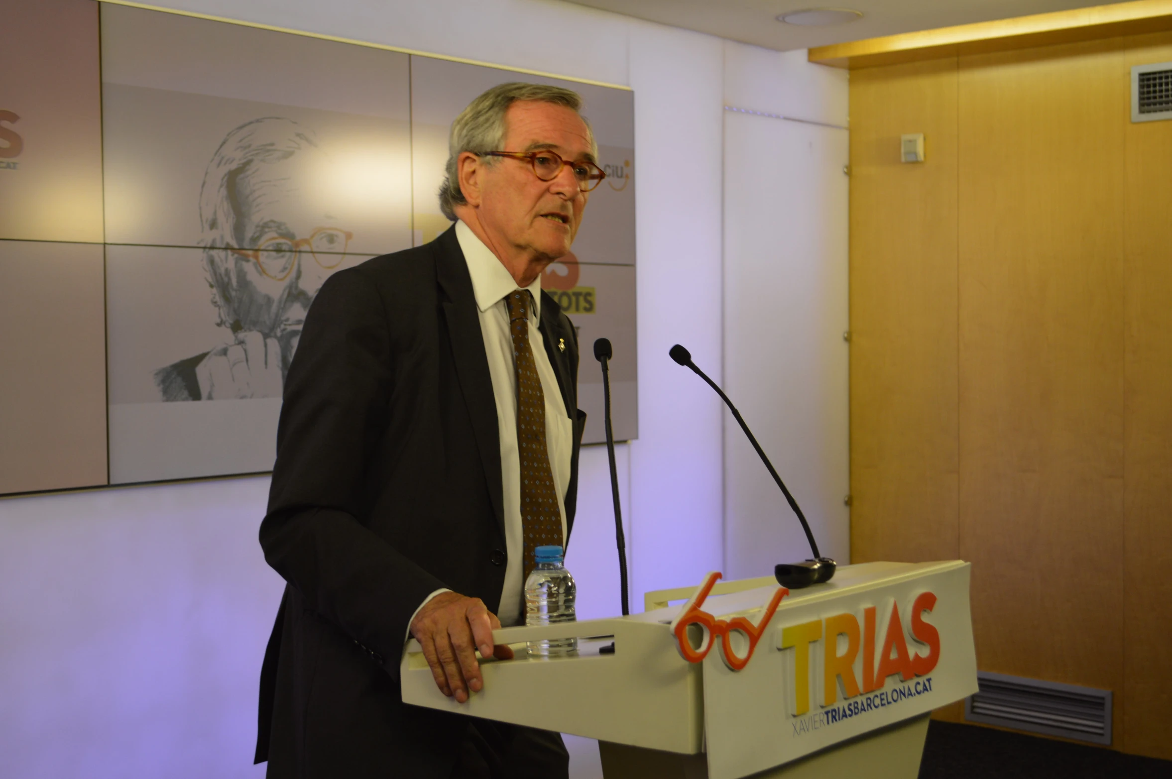 an older man with glasses stands behind a podium while speaking at a public event