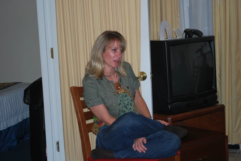 a woman in blue jeans and green shirt sitting on chair with a television behind her