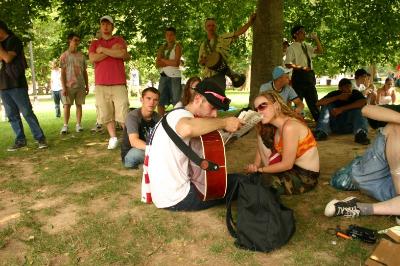 there is people standing and sitting in the grass, one man playing guitar