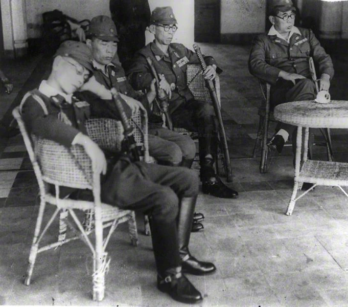 an old black and white pograph shows men in uniforms sitting on chairs, in a room with a table