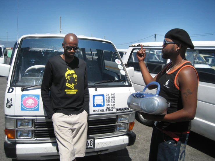 two men stand next to each other in front of parked cars