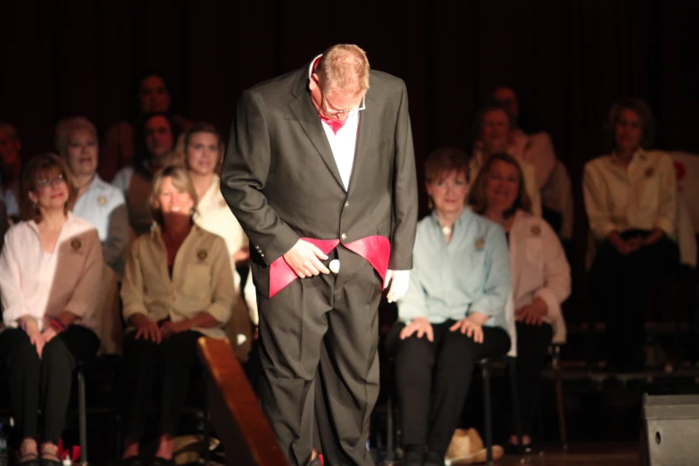 a man with a red tie stands on stage while people sit behind him