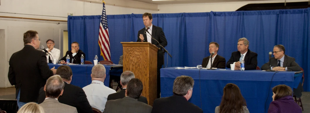 a man giving a speech from a podium with other people sitting around