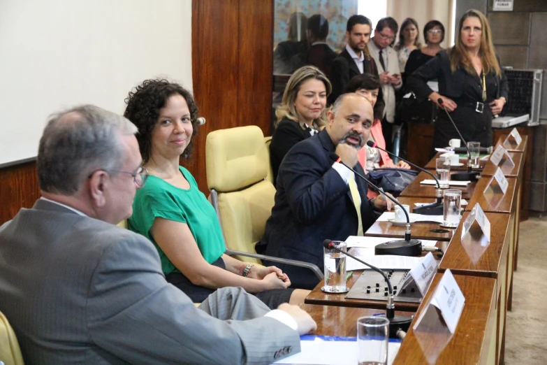 people are watching others at an event, sitting at a long conference table