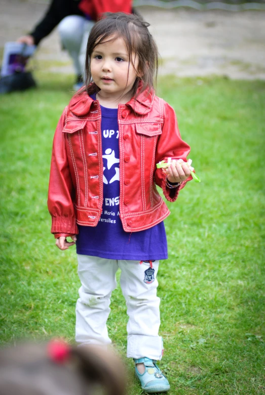 a little girl in red jacket and white pants eating soing