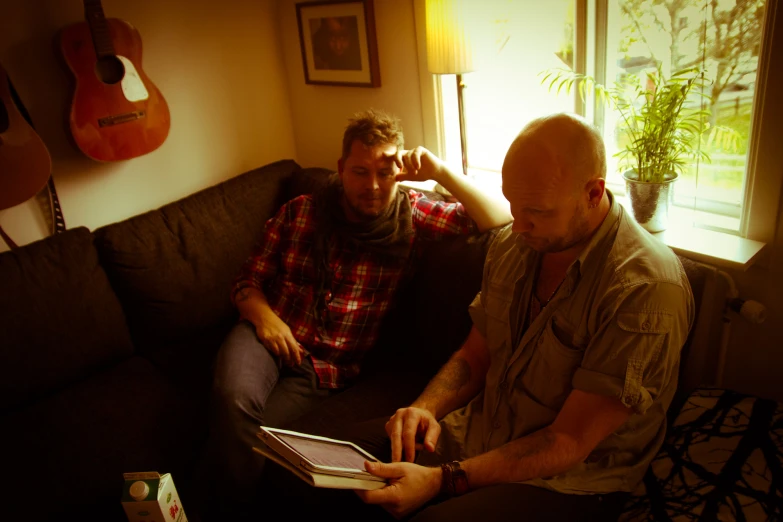 two men sit on a couch while reading a book