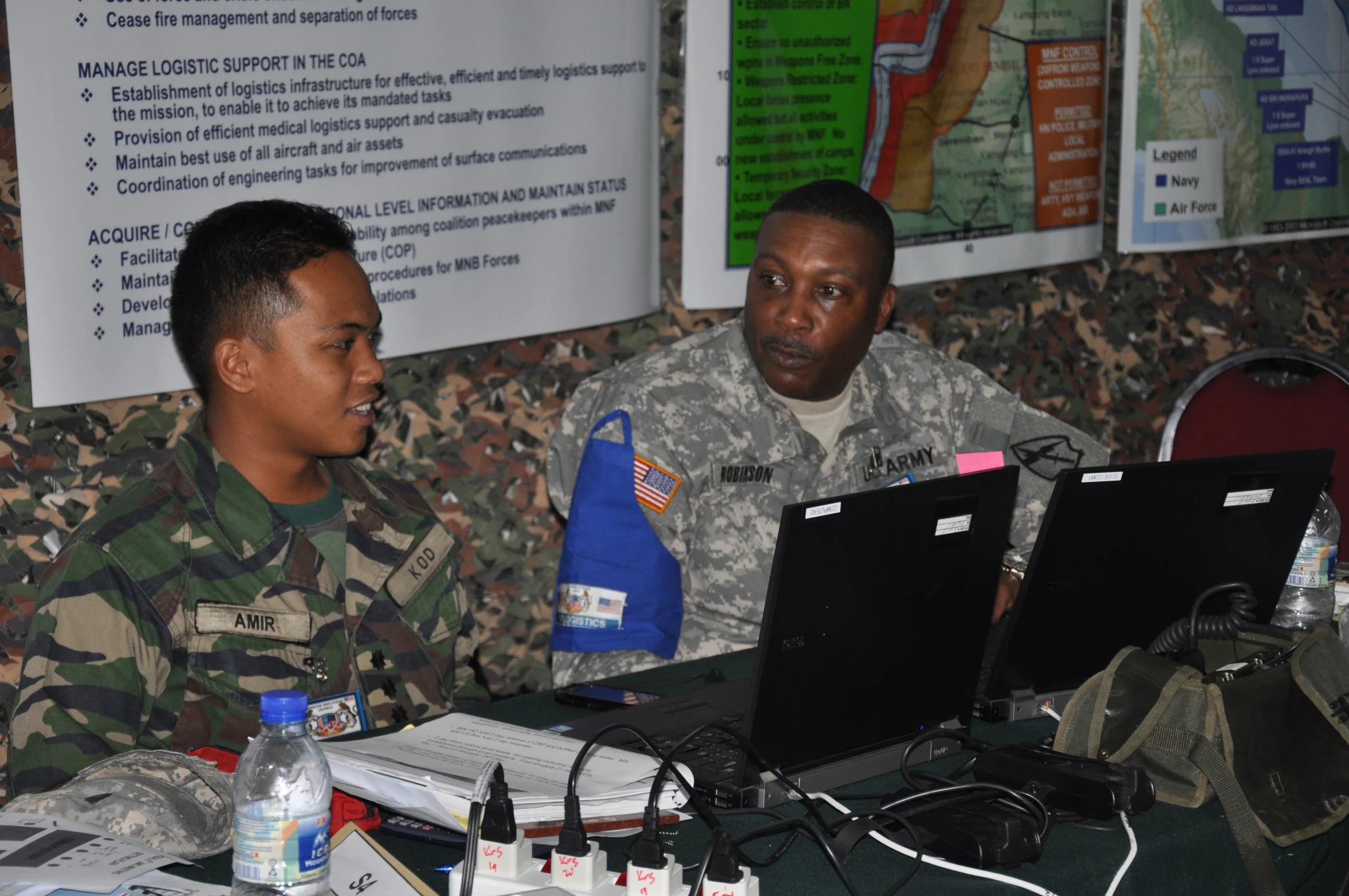 two military men sit and talk in front of laptops