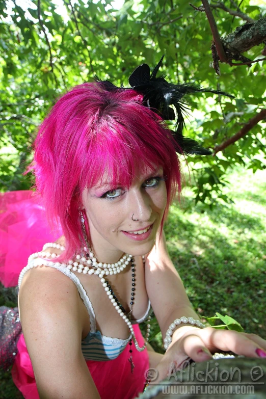 a woman with bright pink hair and pearls sitting on a grass covered park