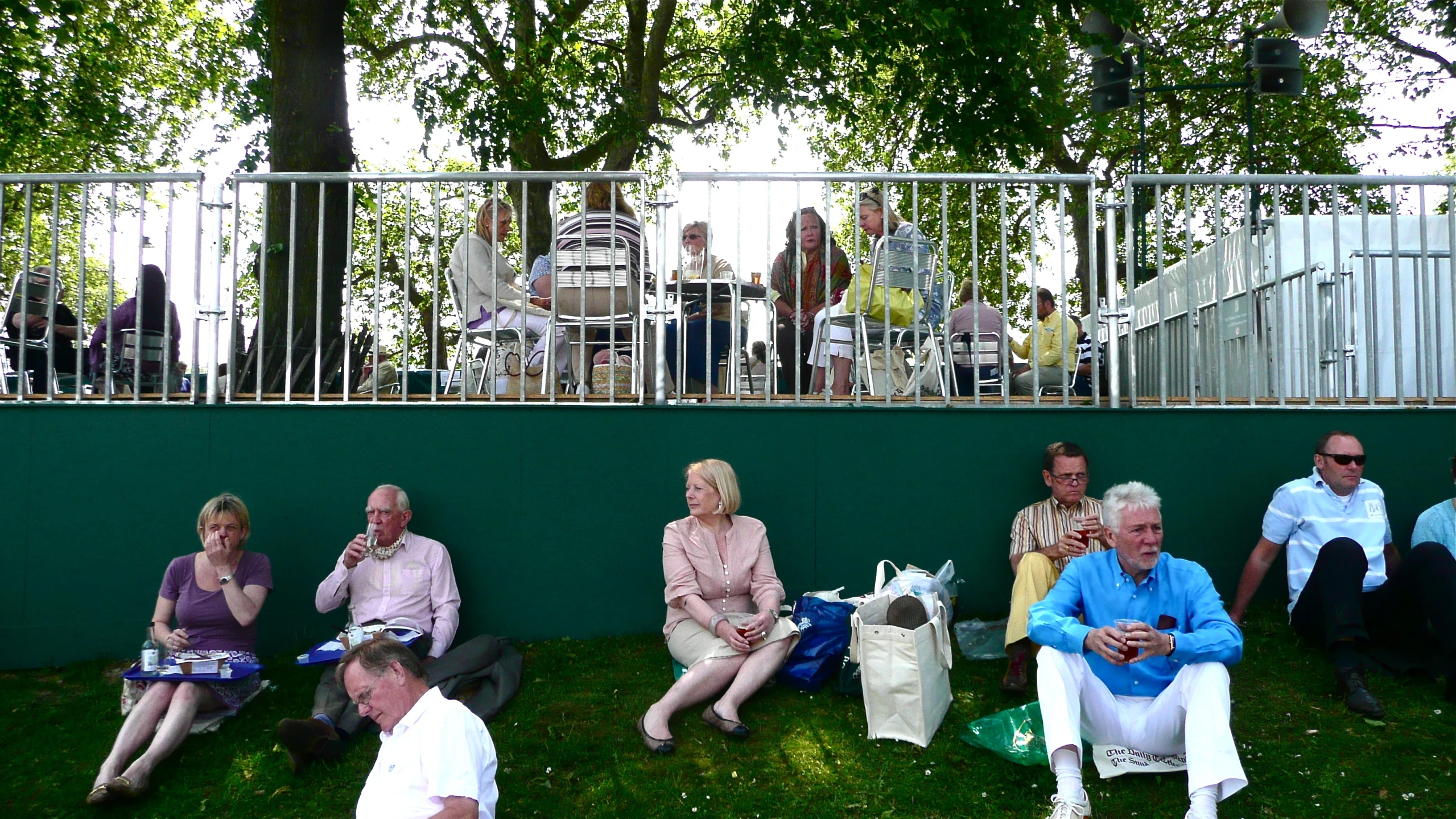 several people are sitting on the grass beside a green wall