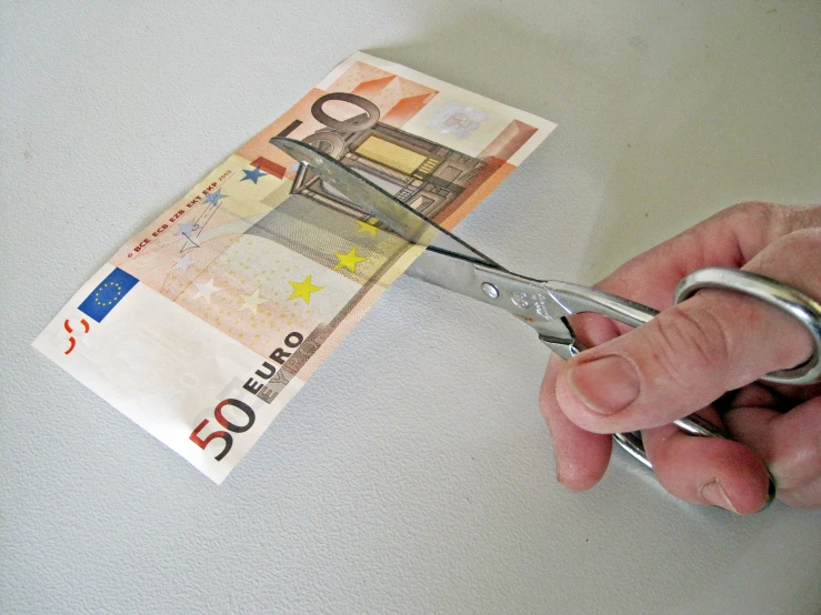a hand holding a pair of scissors with a bank note below it