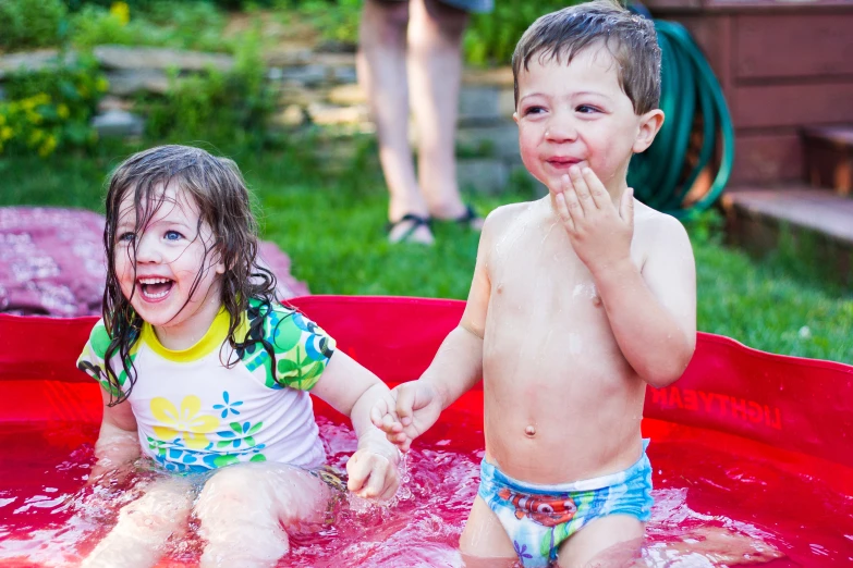 two children, one sitting in a pool one is smiling at the camera while one holds his hand over his mouth