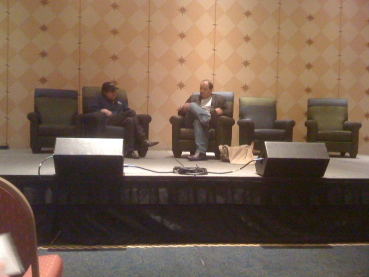 three men sitting on the stage one of them is listening to another