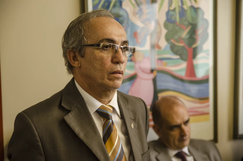 two men sitting in front of paintings, one of them is wearing a suit