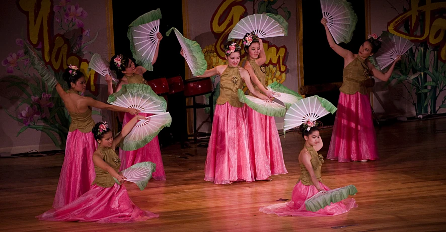 a group of women in costume stand in front of two small women holding large white fans