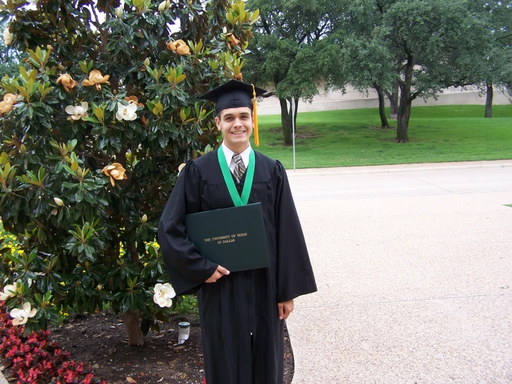 a man standing in front of a tree wearing graduation gown and holding an award