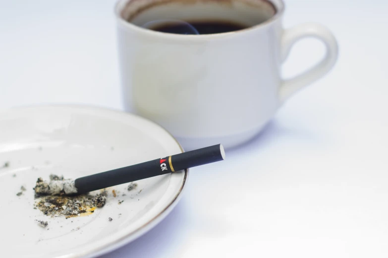 a cigarette resting next to a plate with cigarettes next to a cup of coffee