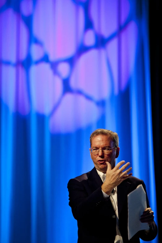 a man in black jacket holding a laptop with a blue curtain behind him