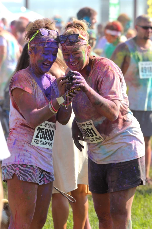 two women covered in colored mud are on their phones