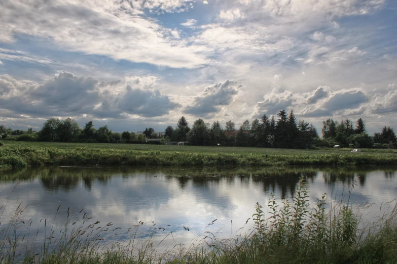 some clouds are floating over a pond with trees and grass