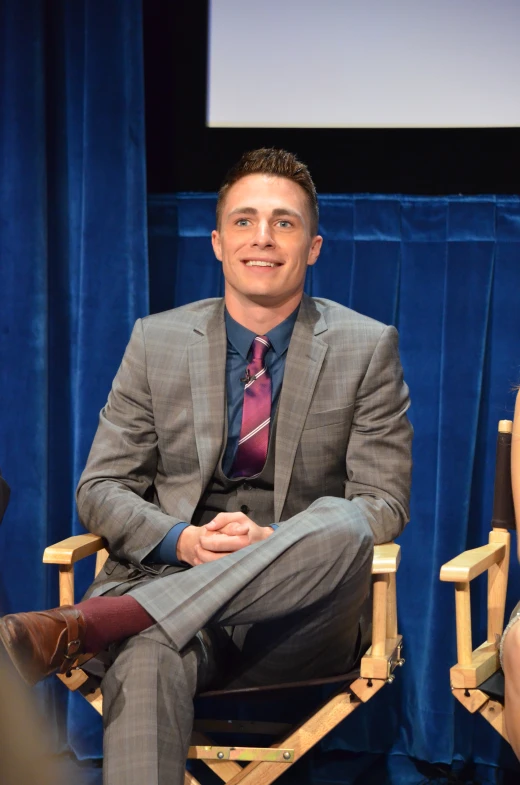 a man sitting down on a wooden chair in front of a blue curtain