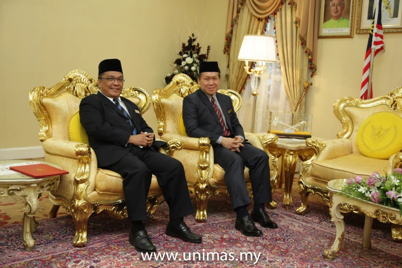 two men sit in gold chairs together in a living room