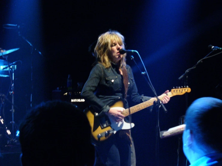 a girl in black shirt holding a yellow guitar and singing at microphone