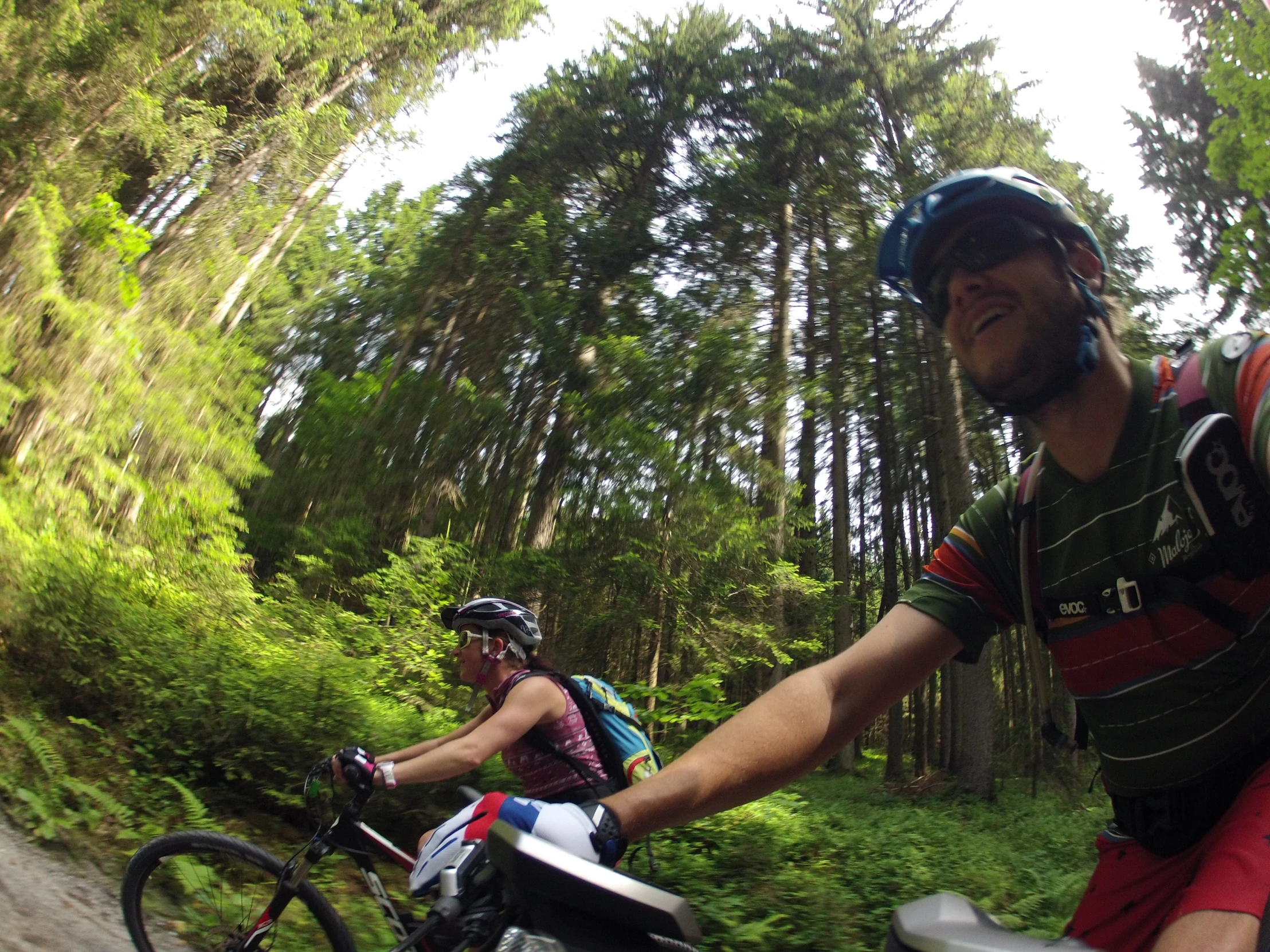 two people wearing helmets riding bikes down a forest