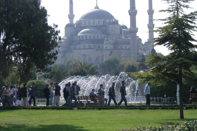 a number of people walking past a fountain with a large building in the background