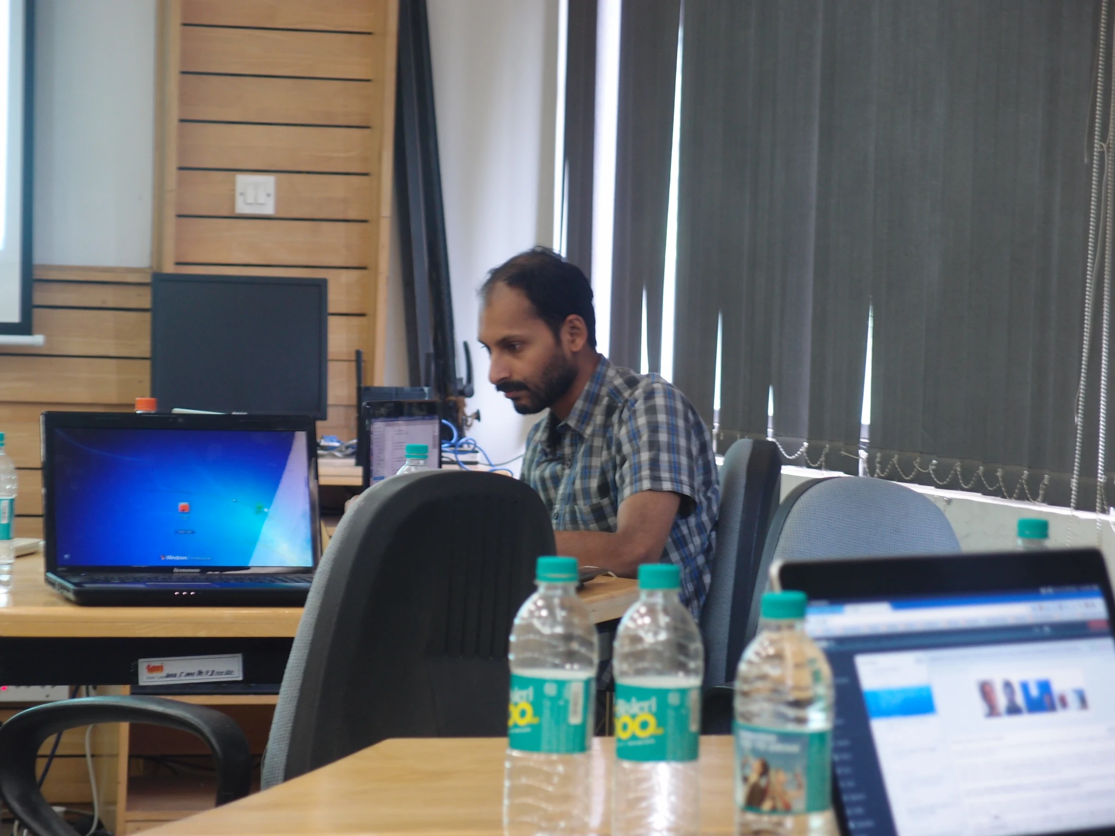 man in plaid shirt at desk with computers and water bottles