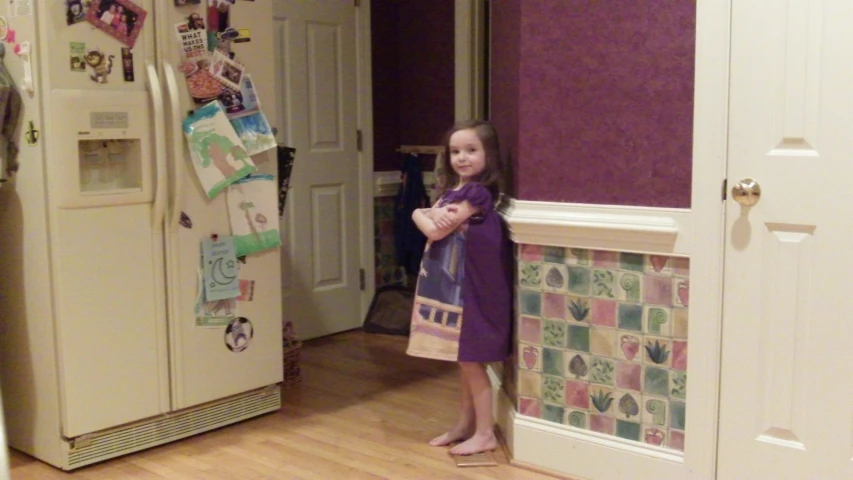 a little girl standing by a door and a refrigerator