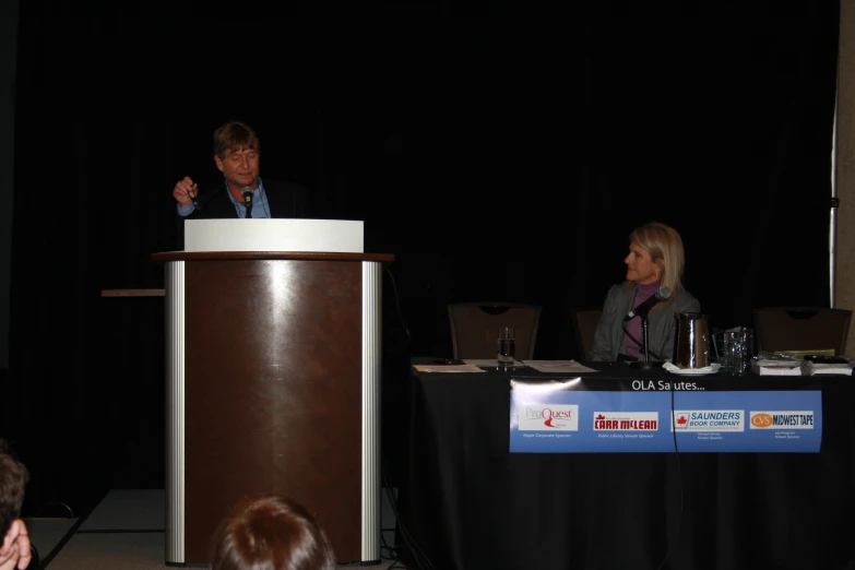 a woman talking at a podium in front of a man