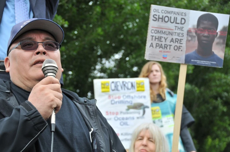 man speaking into a microphone next to people in front of trees