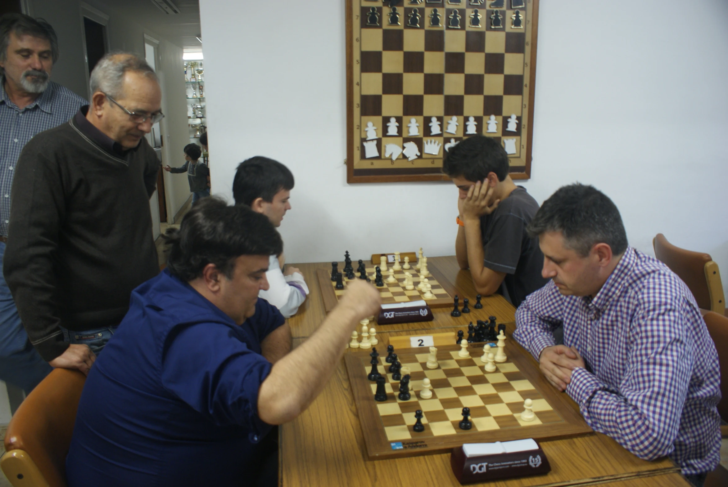 an elderly man standing next to two younger men playing chess