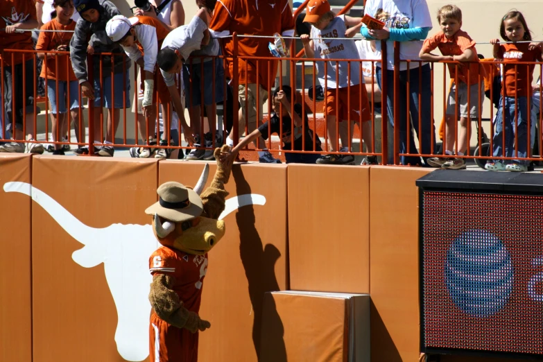 a scarecrow in an orange and white hat is next to a fence and there is a crowd of people behind a barrier