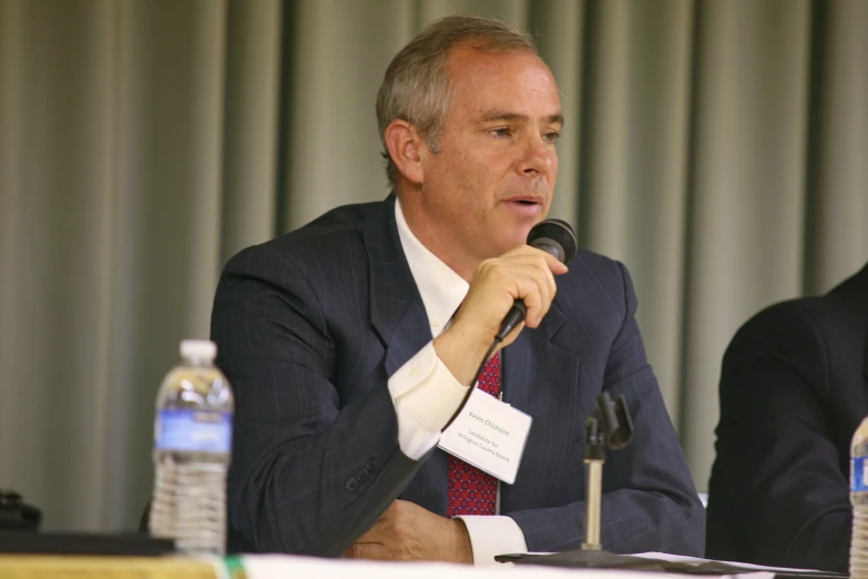 a man in suit and tie sitting at a table while talking into a microphone