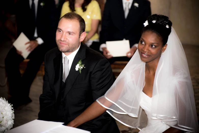 a couple in wedding attire sitting at a priest's desk