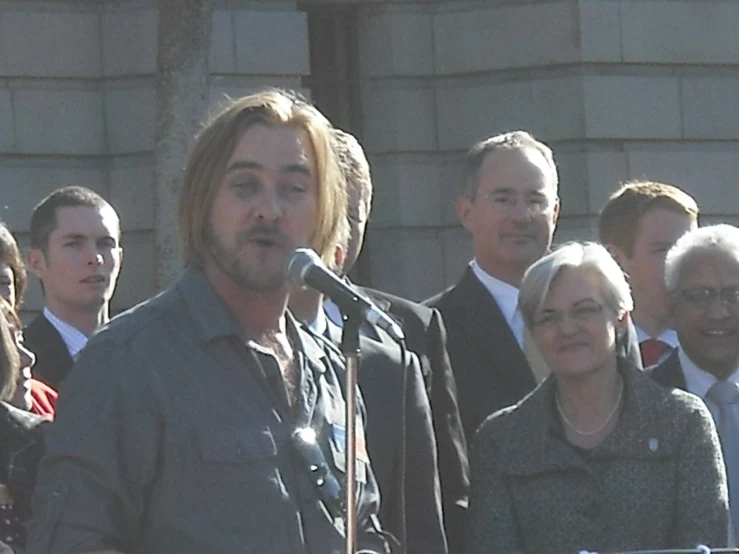an image of man speaking into microphones at outdoor event