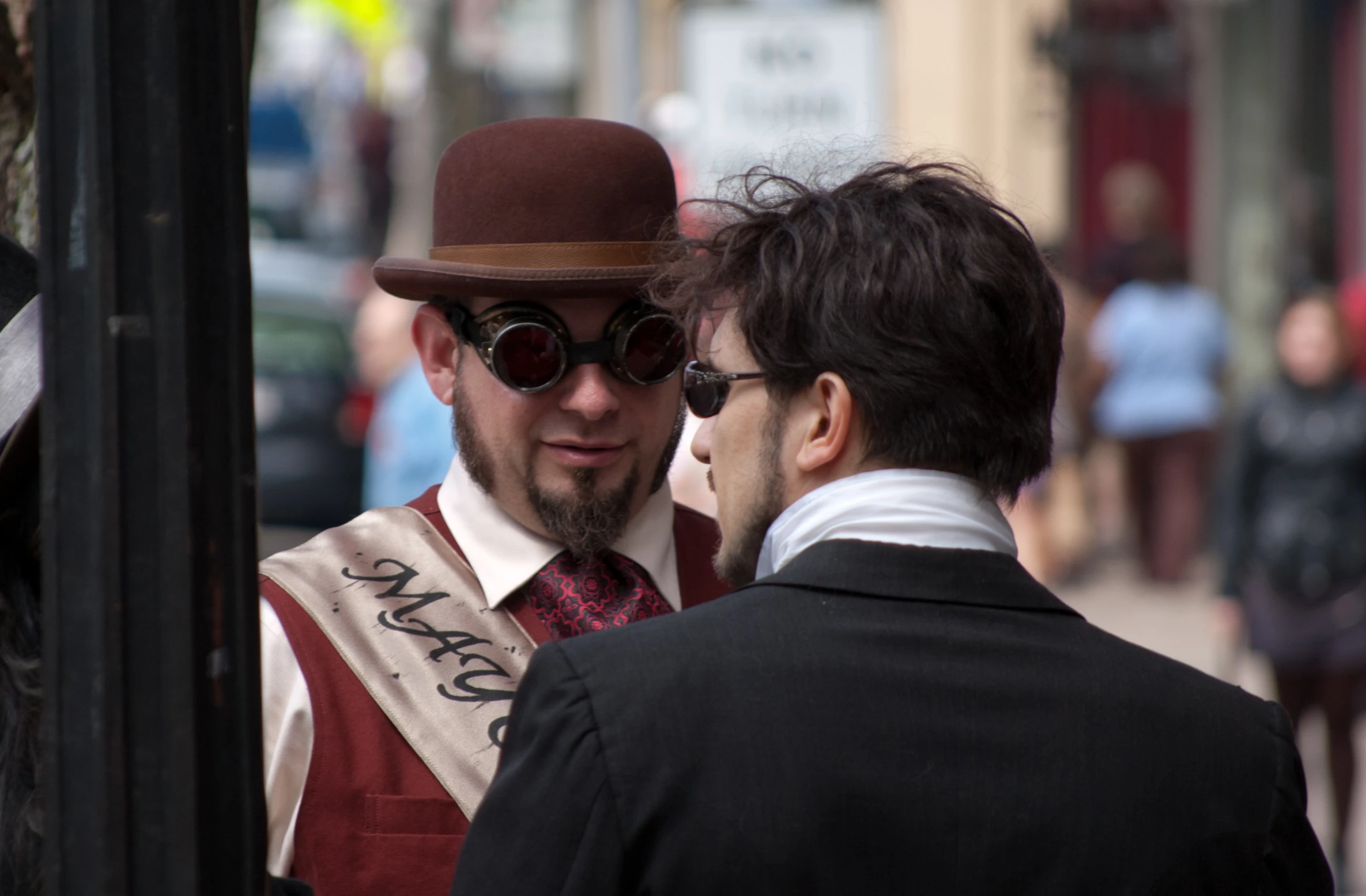 a man in an old fashioned suit and bowler hat talking to another man on the street