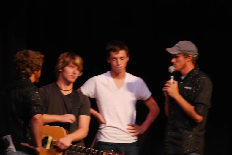 three young men, one with a guitar and the other a microphone, standing in front of a dark backdrop