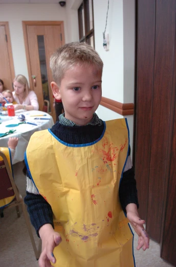 a boy wearing an apron stands in a room full of other children